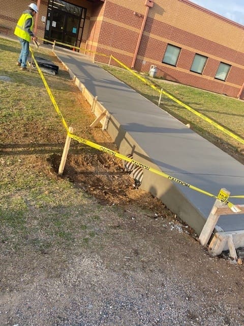 Construction worker in a high-visibility vest smoothing freshly poured concrete on a sidewalk next to a brick building, with caution tape around the site.