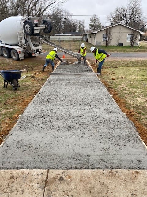 Three construction workers in safety vests and helmets smoothing freshly poured concrete from a mixer truck to form a sidewalk.
