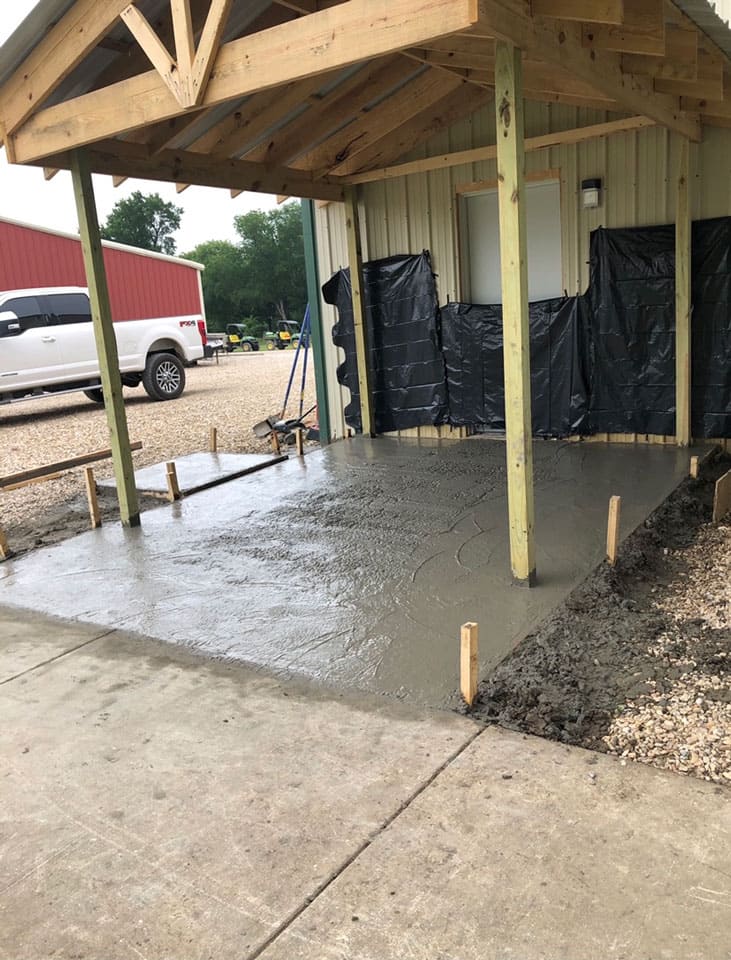 Newly poured concrete slab under a wooden shelter attached to a metal-sided building, with a white pickup truck and red barn in the background.