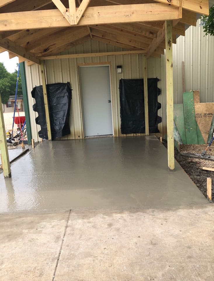 Newly poured smooth concrete slab under a wooden roof structure with a closed door at the back wall of a metal building.