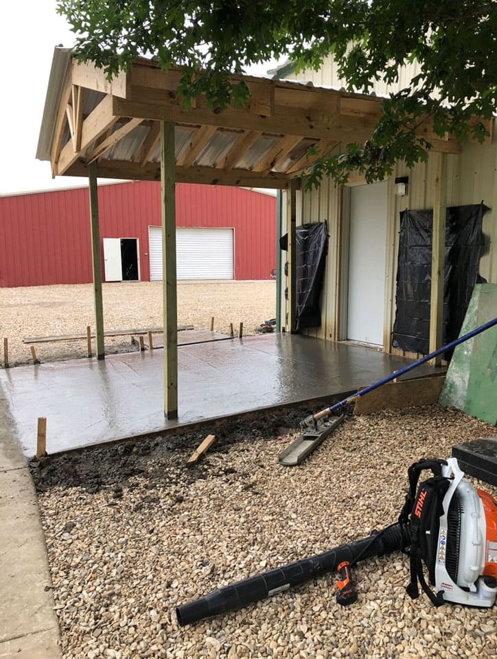 Newly poured concrete slab under a wooden canopy attached to a building, with a leaf blower and landscaping rake on gravel in the foreground.