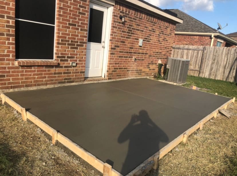 Newly poured smooth concrete slab patio next to a brick house with a white door and window.