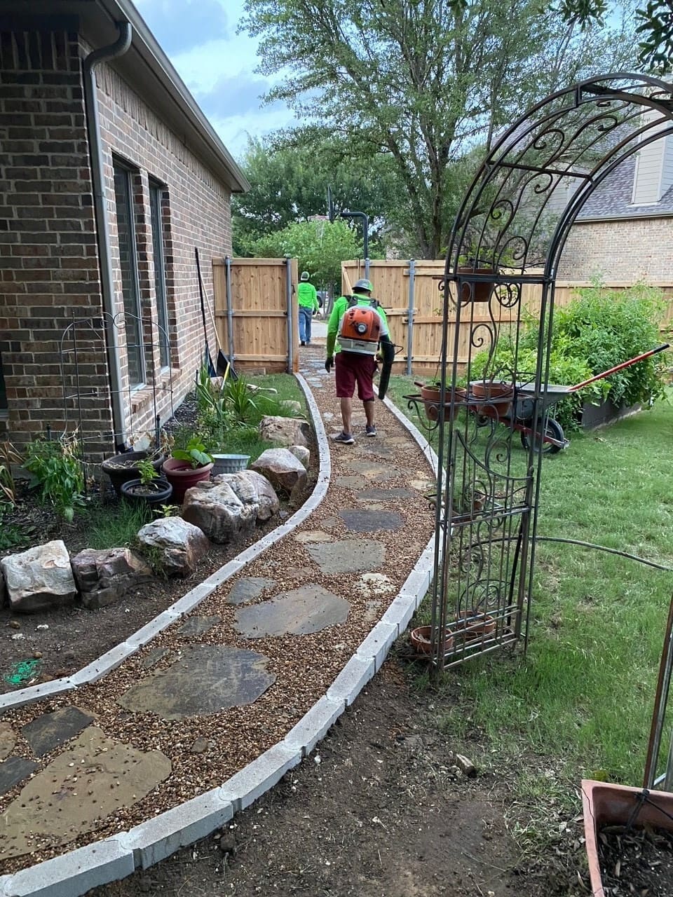 Two workers in bright green shirts walking on a gravel and stone garden path beside a brick house, with a metal garden arch and garden tools nearby.
