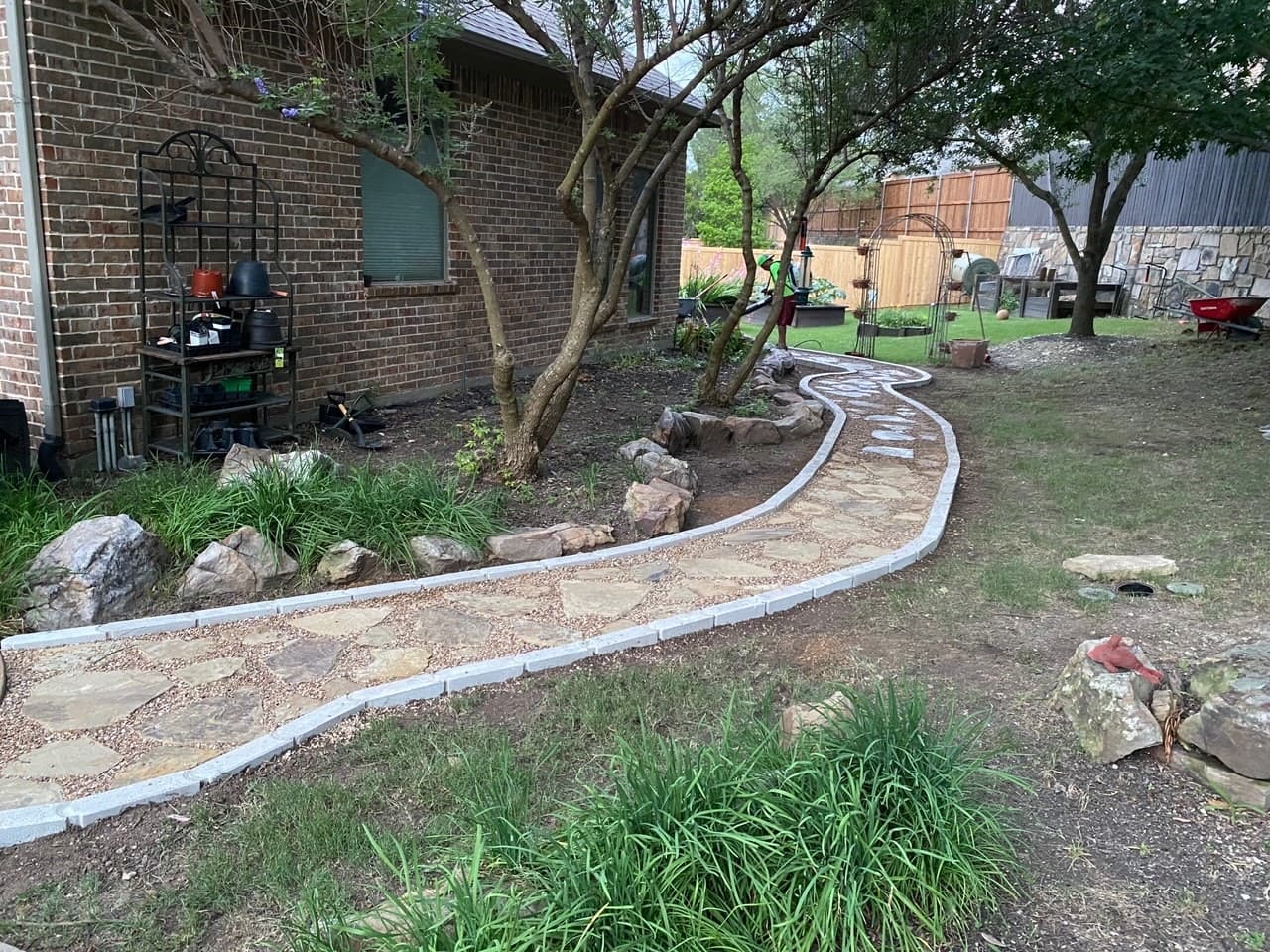 Stone pathway lined with bricks winding through a backyard with trees and garden beds beside a brick house.