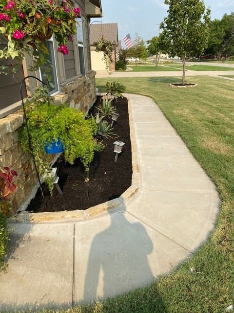 A curved concrete sidewalk next to a garden bed with dark mulch, small plants, a hanging green plant in a blue pot, and a house with stone and siding exterior.