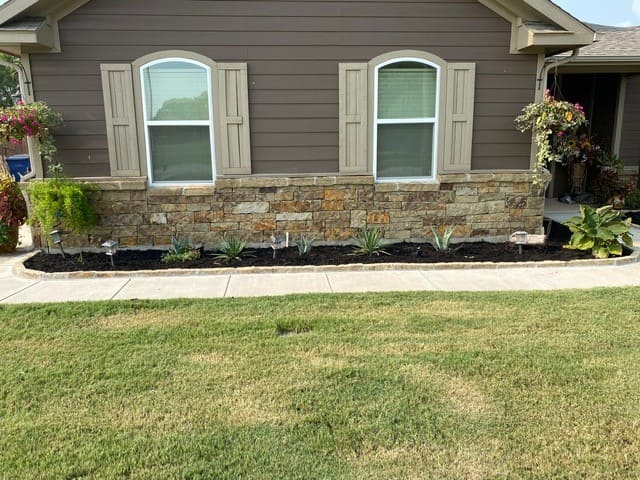 House exterior with brown siding, two white-trimmed windows above a stone facade, a flower bed with small plants, and a green lawn in front.