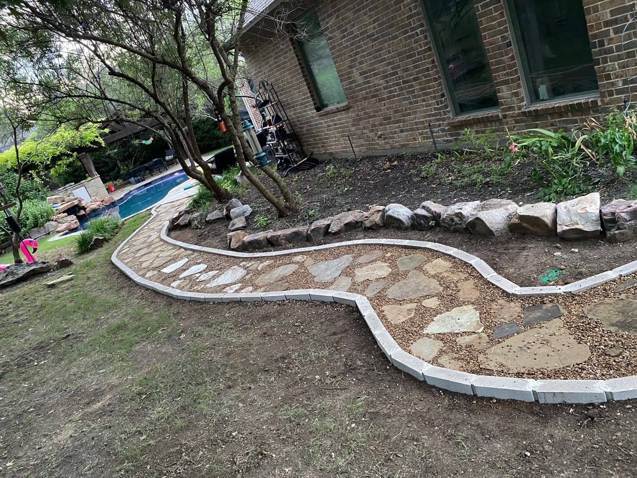 Curved stone and gravel pathway bordered with pavers alongside a brick house and yard with trees and a pool in the background.