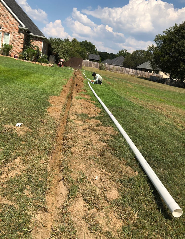 Person working on installing a long white pipe beside a freshly dug trench in a grassy yard near a brick house.