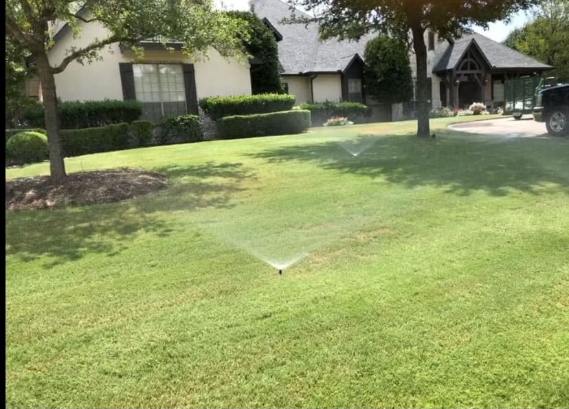 Green lawn with two sprinklers watering the grass in front of a house with trees and shrubs.