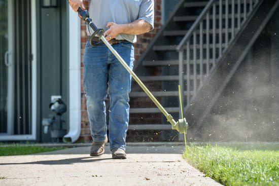 Person using a string trimmer to cut grass along a sidewalk edge near stairs and a building.