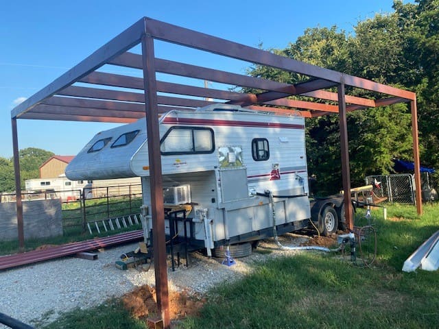 White camper trailer parked under a partially constructed metal frame shelter on a grassy and gravel area.