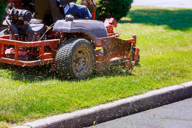 Close-up of a person mowing grass with a red riding lawn mower near a sidewalk curb.