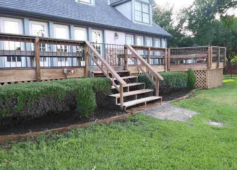 Wooden deck with stairs and railing attached to a two-story house with multiple windows and trimmed bushes along the deck.