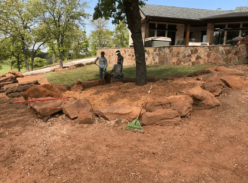 Three people working on landscaping around a tree with rocks and mulch near a stone wall and house.