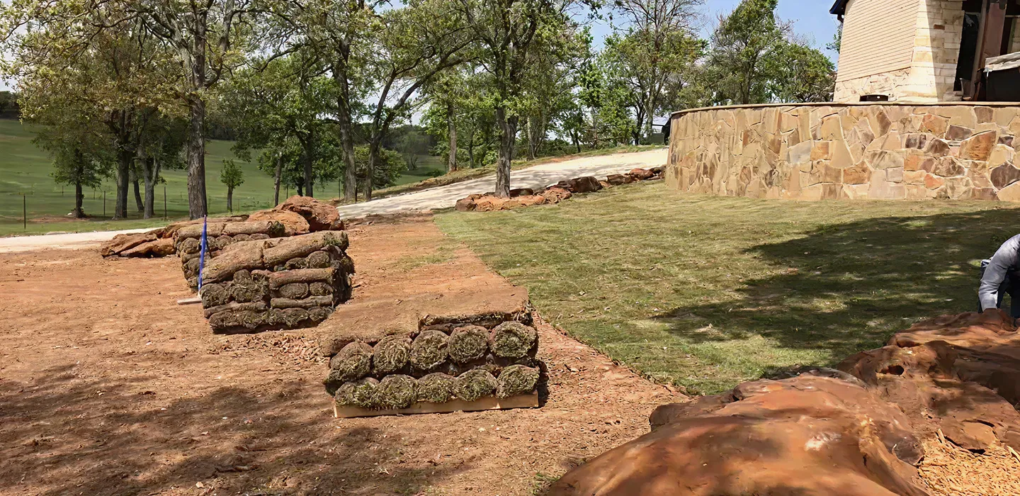 Stacks of rolled sod placed on bare soil near a stone wall and grassy area with trees in the background on a sunny day.