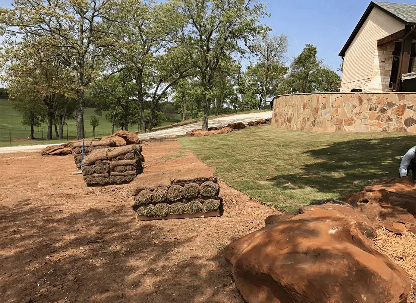 Stacks of rolled sod on a dirt area next to a newly laid grassy yard and a house with a stone retaining wall.