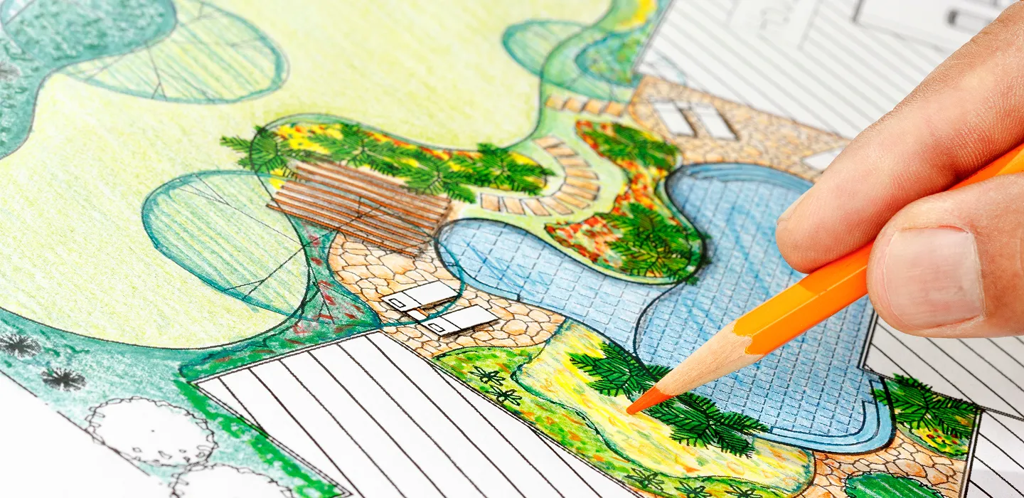Hand holding an orange pencil coloring a detailed landscape design plan with water features, pathways, and greenery.