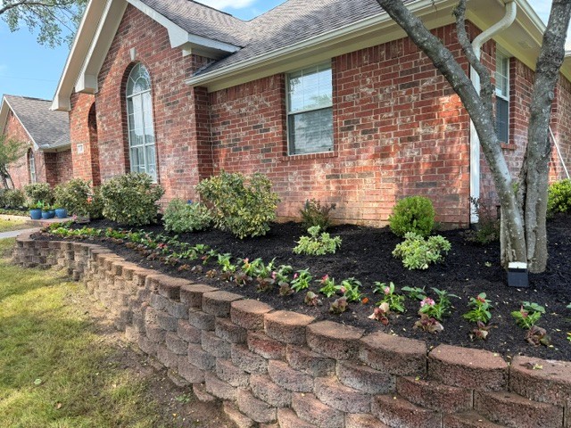 Brick raised garden bed with small plants in front of a brick house under a blue sky.